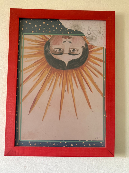 A framed art print featuring a stylized sun with a human face, displayed on a textured off-white wall. The print has a red wooden frame and depicts a celestial design with rays, stars, and a dark green night sky.